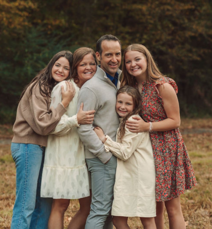 Family portrait: Dad surrounded by his wife and three daughters, embracing outdoors.